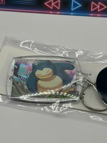 A Snorlax character keychain in a clear plastic protective sleeve, featuring a rectangular card with an illustration of Snorlax and a silver metal key ring.