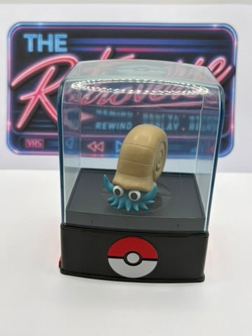 A small figurine of a snail-like Pokémon with a tan shell and blue tentacles, displayed inside a clear acrylic case on a black base with a Pokéball logo. A neon-style sign reading 'THE Rewind' is visible in the background.