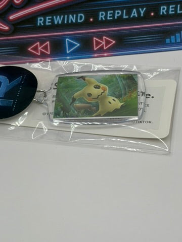 A packaged rectangular keychain featuring a mimikyu, shown with a round black keychain charm and a white card insert on a white surface. A music player interface graphic is visible at the top of the frame.