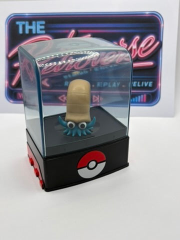 A small collectible figurine displayed inside a clear acrylic dome on a black base with a red-and-white Pokéball logo, set against a neon-style sign and white background.