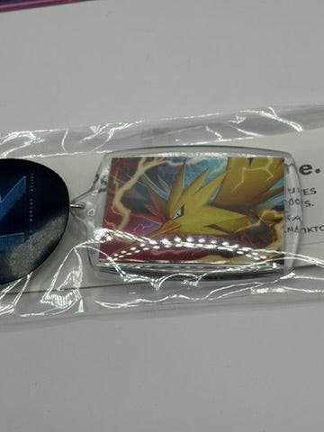 A Pokémon keyring featuring a colorful illustration of Zapdos, sealed in clear plastic packaging on a light gray surface.