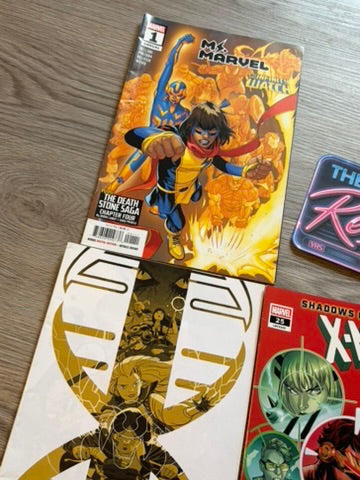 Several Marvel comic books and a card are arranged on a light wood floor. The most prominent item is a comic book titled 'Ms. Marvel