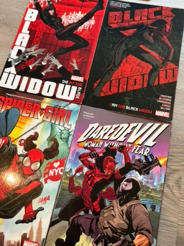 Four Marvel comic books laid out on a light wood surface: Black Widow issue 5, Spider-Gwen issue 2, Daredevil issue 1, and an issue featuring Spider-Gwen and Black Widow together.