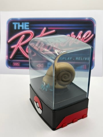 A Pokémon Omanyte figurine inside a clear display case mounted on a black and red base, with a neon-style sign in the background.