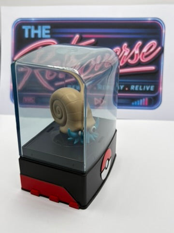 A Pokémon Omanyte figurine inside a clear dome display case with a black and red base, set against a white background with a neon sign behind it.