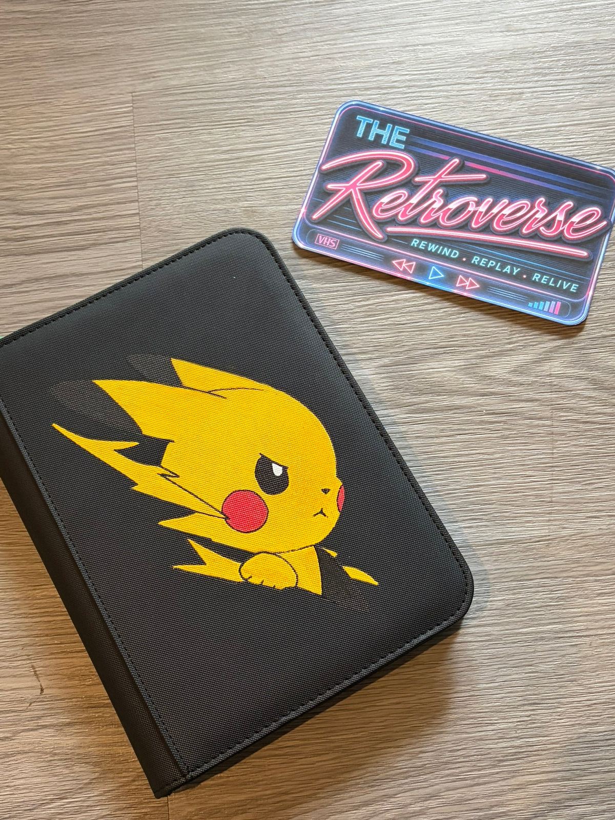 A black faux-leather zippered case featuring a printed Pikachu design, shown on a light wood floor next to a retro neon-style sticker that reads 'THE Retroverse'.