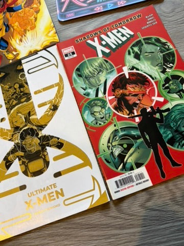 Two X-Men comic books laid on a wooden surface: the right one is Shadow of Tomorrow #2 with a red cover and green/black character art, and the left one is Ultimate X-Men with a white and gold cover.