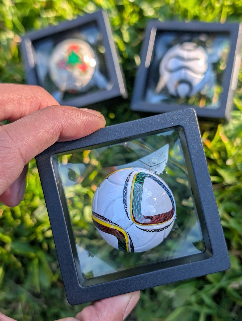 Hand holding a small soccer ball in a clear display case with grass in the background